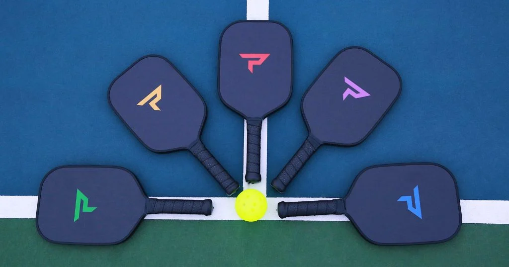 Năm cây vợt Paddletek mặt đen, logo năm màu, xếp hình quạt quanh quả bóng pickleball trên sân xanh