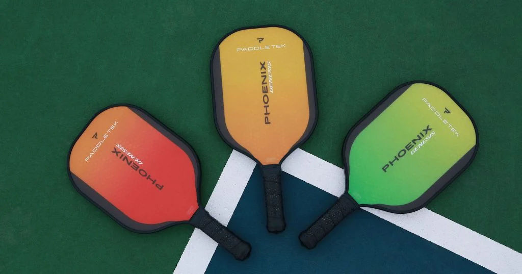 Ba cây vợt Phoenix Genesis màu đỏ, cam vàng và xanh lá đặt trên vạch sân pickleball